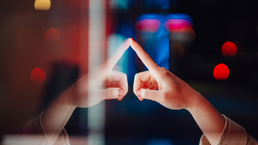 A single hand reaches out to touch its own reflection on a glass surface, with colorful out‑of‑focus lights creating a glowing backdrop.