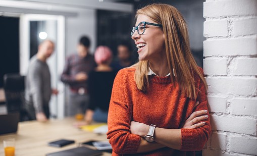 Person in orange sweater standing in front of a white brick wall; blurred office scene in background.