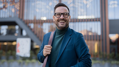 Person in blue blazer and green turtleneck standing in front of a modern building with wooden slats.