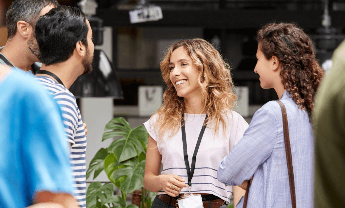 A group of four people stand in a circle, engaged in conversation. A green plant and hanging lights are in the background.
