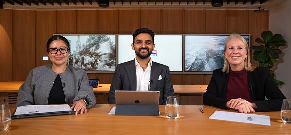 Three people sit at a table on a talk show set with documents and a laptop, three large display screens in the background.