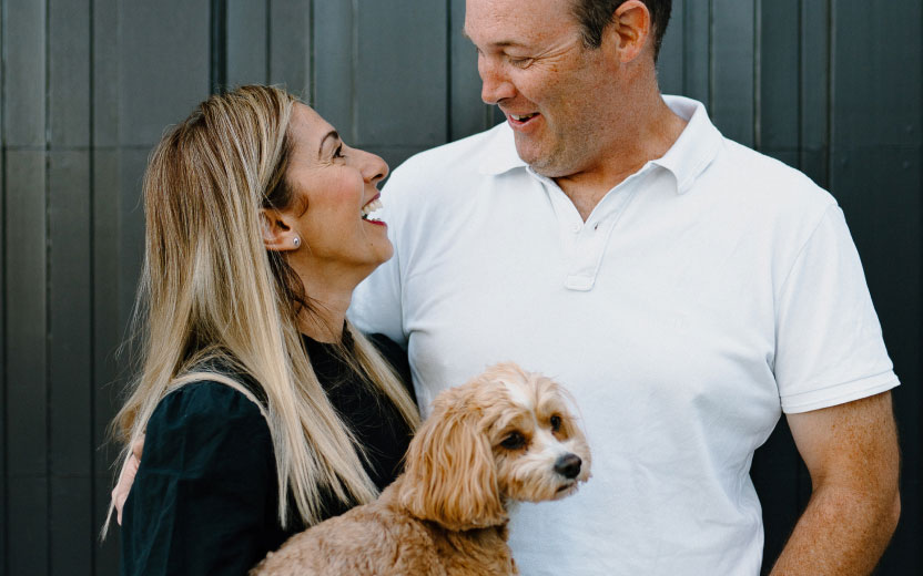 A couple standing together outdoors while holding a small dog in their arms.