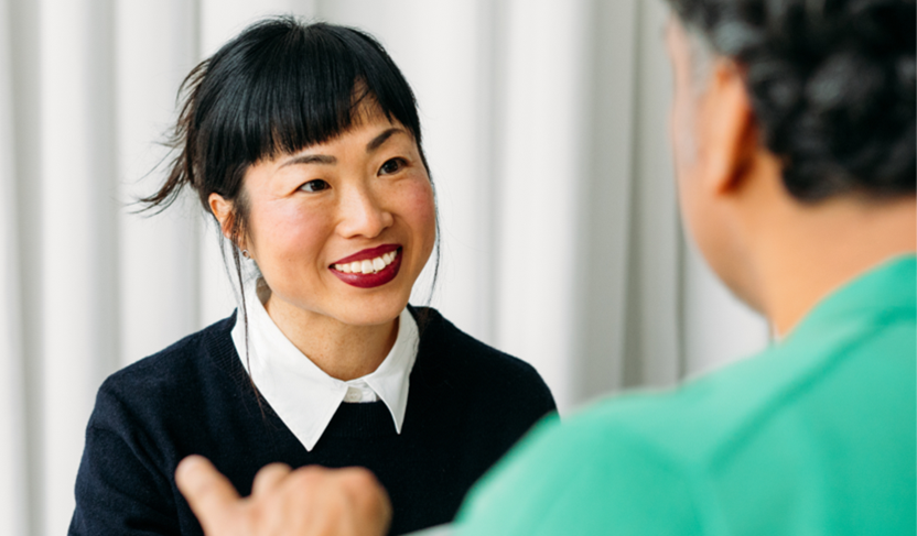 Two people sitting indoors having a conversation, with one facing the camera and the other turned away.