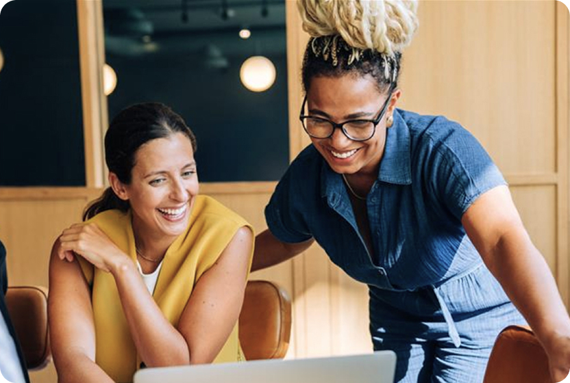 Two people are working together at a table while looking at a laptop screen.