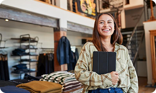 A person standing in a clothing store, holding a tablet against their chest, with shelves of folded clothes and hanging garments in the background.